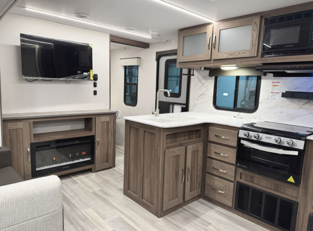 Interior of an RV kitchen and living area featuring wood cabinetry, a white marble-look countertop, and a stainless steel sink with a high-arc faucet. The kitchen includes a black stovetop and oven, a microwave, and a window above the counter. Adjacent to the kitchen is an entertainment center with a mounted flat-screen TV and an electric fireplace below. The flooring is light wood-look vinyl, and the ceiling has recessed LED lighting.