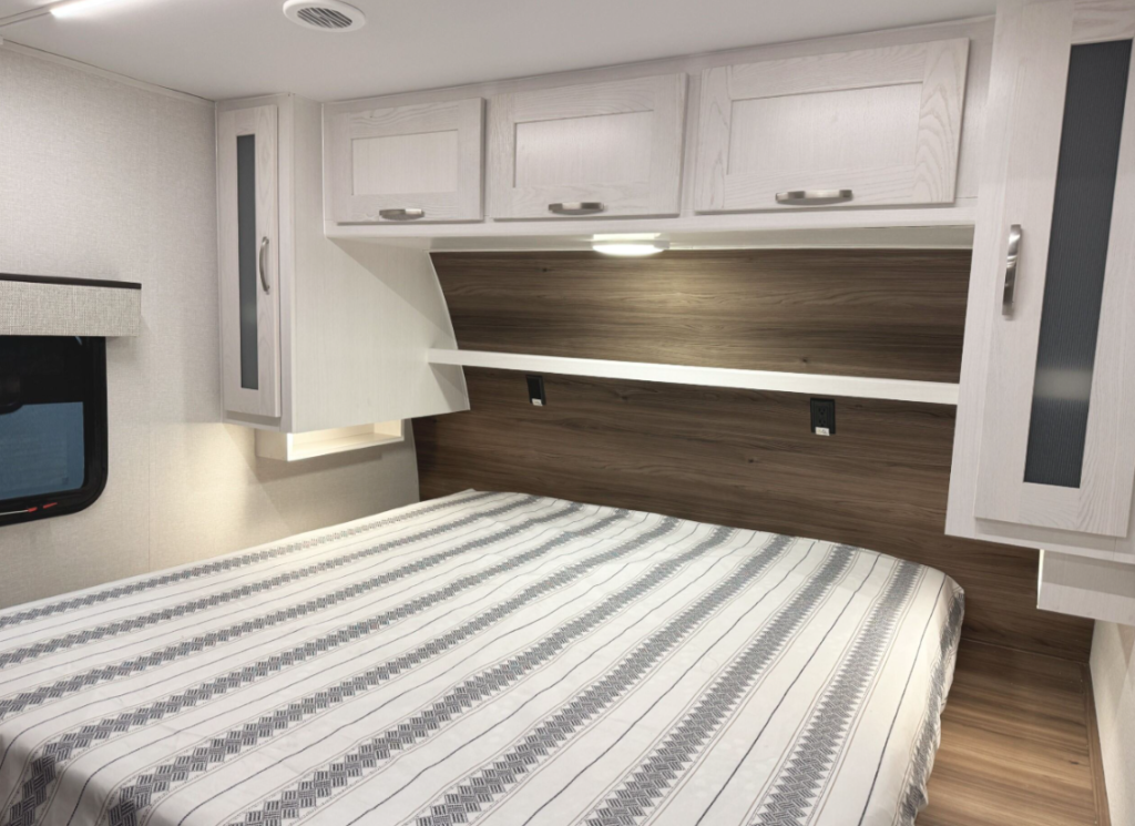 RV bedroom featuring a queen-size bed with a striped gray and white bedspread. Above the bed, there are white woodgrain cabinets with silver handles for additional storage, along with a built-in shelf and reading light. The headboard area is accented with warm wood paneling. A small window with a fabric valance is on the left side, providing natural light. The design combines light and dark wood tones for a modern, cozy feel.