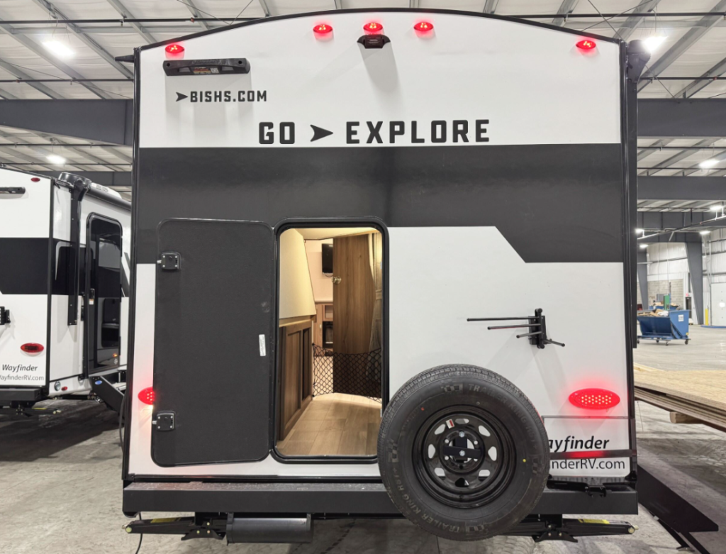 Rear view of an RV with a black and white exterior featuring the slogan "GO > EXPLORE" and the website "BISHS.COM" displayed on the back. The RV has a spare tire mounted on the lower section, red brake lights, and a black rear storage access door that is open, revealing a portion of the interior with wood accents and netted storage. A TV mount is installed on the back wall. The RV is parked inside a large, well-lit warehouse alongside other RVs.