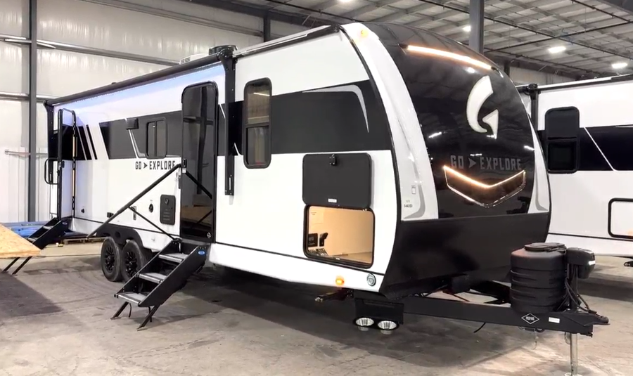  Modern travel trailer with a sleek black and white exterior, featuring the "GO EXPLORE" branding on the front. The trailer has an aerodynamic front cap with integrated LED accent lighting. A large storage compartment is open on the side, and the entrance features fold-out metal steps with a sturdy handrail. The dual-axle RV is parked inside a spacious warehouse alongside other trailers. A power tongue jack and propane tanks are mounted at the front, enhancing convenience for towing and setup.