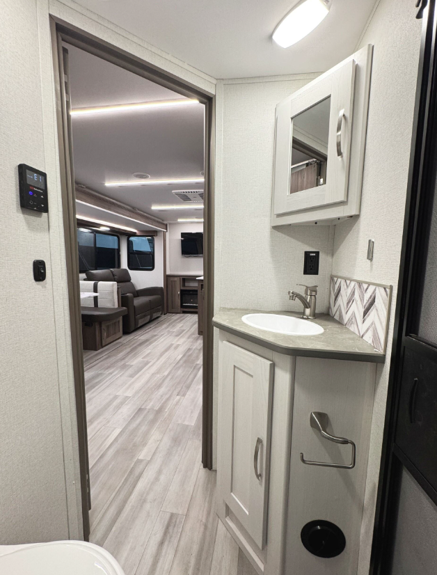  RV bathroom with a compact vanity featuring a light gray cabinet, a marble-look countertop, and a small round sink with a brushed nickel faucet. Above the sink is a mirrored medicine cabinet, and a modern geometric tile backsplash adds a decorative touch. A black outlet and a stainless steel towel ring are mounted on the wall. The flooring is light wood-look vinyl. The view extends into the main living area, which includes a brown leather loveseat, a dinette, and an entertainment center with an electric fireplace and a mounted TV. LED strip lighting runs along the ceiling, enhancing the modern and spacious design.