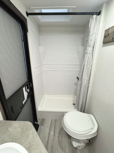 RV bathroom featuring a white shower with a textured subway tile pattern and a decorative trim. A striped shower curtain hangs from a black rod, and a skylight above allows natural light. The space includes a compact white toilet and a small vanity with a marble-look countertop. A black-framed door with a built-in screen is on the left side, providing ventilation and access to the exterior. The flooring is light wood-look vinyl, complementing the neutral color scheme.