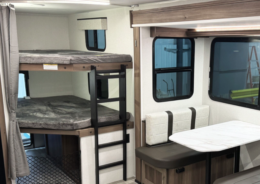 RV interior featuring a bunk bed area with two twin-sized beds, each with plush gray mattresses. A black metal ladder provides access to the top bunk, and a small window with a fabric valance is positioned beside it. A privacy curtain is attached to the side for added separation. Adjacent to the bunk beds is a dinette with a white marble-look table and cushioned bench seating in brown and cream tones. Large windows surround the dinette area, allowing for natural light. The interior design incorporates warm wood tones and neutral colors.
