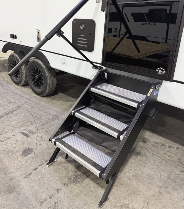 Close-up view of a fold-out aluminum entry step on a travel trailer. The sturdy three-step design features anti-slip treads and adjustable legs for stability on uneven surfaces. A black entry door with a reflective window is visible, along with a grab handle for added support. The RV’s exterior includes a white and black color scheme, a storage compartment, and a dual-axle setup with black alloy wheels. The trailer is parked inside a warehouse setting.