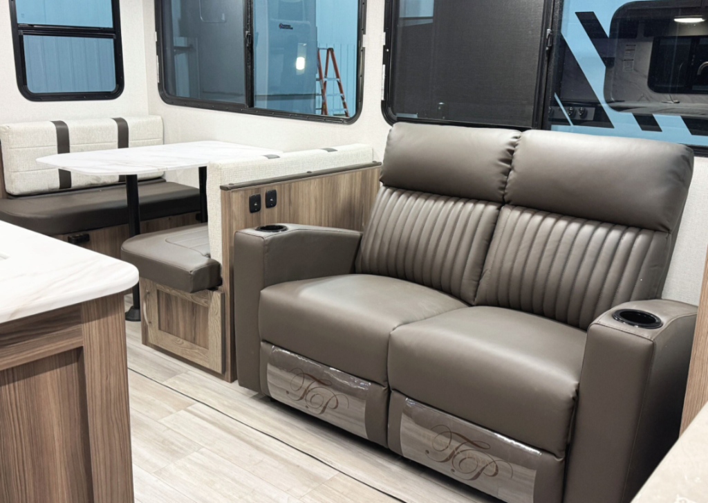 Interior of an RV featuring a brown leather loveseat with cupholders on each armrest and a subtle monogram on the footrests. Adjacent to the loveseat is a dining booth with a white marble-look table and cushioned bench seating in brown and cream tones. The interior has wood paneling, light flooring, and large windows that provide natural light.