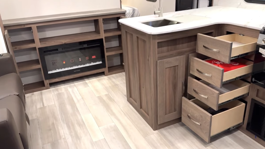 RV interior featuring an open kitchen and living area with wood cabinetry and light wood-look flooring. The kitchen includes a white marble-look countertop with a stainless steel sink and multiple open drawers, showcasing ample storage space. Adjacent to the kitchen, an entertainment center with an electric fireplace is surrounded by open shelving. A brown leather loveseat is partially visible on the left side of the image.