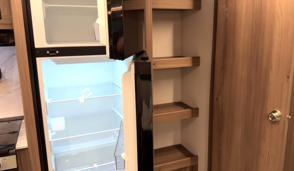 RV kitchen area featuring an open black double-door refrigerator with bright interior lighting and multiple shelves. To the right of the refrigerator, there is a built-in wooden pantry with open shelving for storage. A wooden door with a silver doorknob is visible, leading to another section of the RV. The surrounding cabinetry and walls have a warm wood finish.