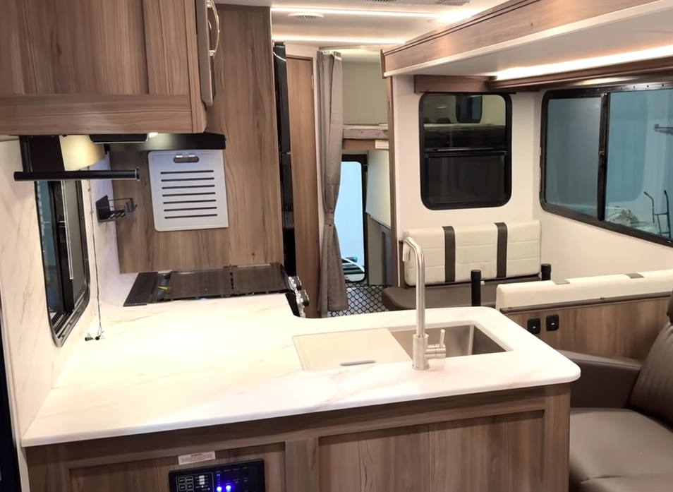 Interior of a modern travel trailer featuring a spacious kitchen with a white marble-look countertop, a stainless steel sink with a high-arc faucet, and a stovetop with a cover. The wood cabinetry and light wood-look flooring create a warm ambiance. In the background, a dinette with cushioned bench seating is next to large windows. A bunk bed area with a privacy curtain and netted storage is visible, along with an open rear door leading outside. LED strip lighting enhances the space, and a brown leather loveseat is partially visible in the foreground.