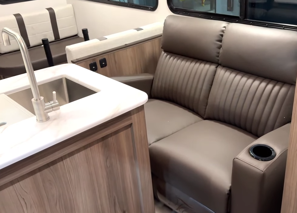 Interior of an RV showcasing a cozy living space with a brown leather loveseat featuring stitched detailing, a built-in cup holder, and plush cushions. The loveseat is positioned next to a kitchen island with a white marble-look countertop and a stainless steel sink with a high-arc faucet. In the background, a dinette with cushioned bench seating in cream and brown tones is situated beside large windows, allowing for natural light. The interior design blends warm wood cabinetry with modern finishes for a stylish and functional layout.