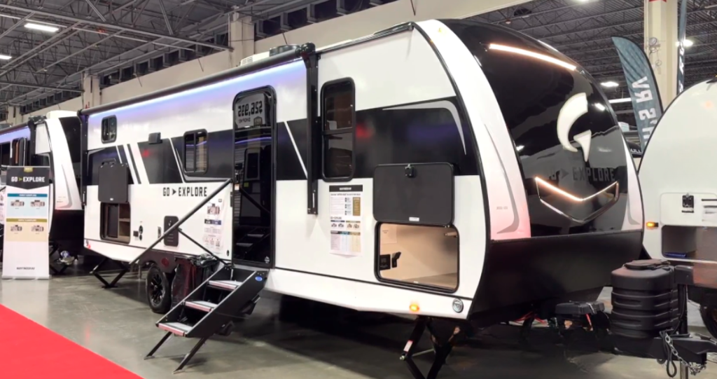 A sleek, modern travel trailer displayed indoors at an RV show. The trailer features a bold black and white exterior with the "GO EXPLORE" logo and a large front window. It has an entry door with fold-down metal steps, storage compartments, and bright LED lighting along the roofline. Other RVs are visible in the background alongside a red carpet walkway.