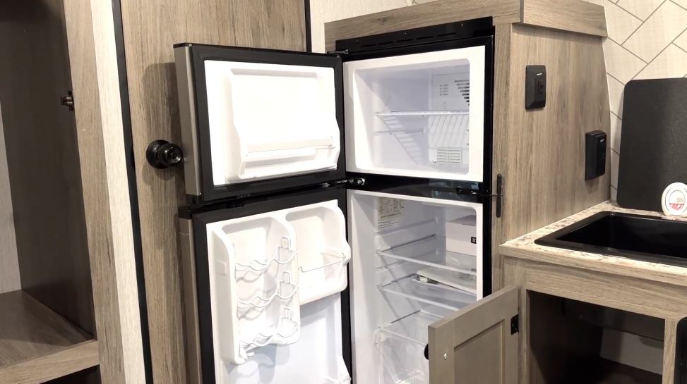 Interior of an RV kitchen featuring an open two-door refrigerator with a separate freezer compartment. The refrigerator has multiple shelves, door storage, and a spacious interior. It is built into a wood-paneled cabinet that matches the surrounding light wood decor. To the right, a small kitchenette includes a black sink with a cover, a light-colored countertop, and a partially open lower cabinet for additional storage. The backsplash features a modern white subway tile pattern. A doorway in the background leads to another section of the RV.