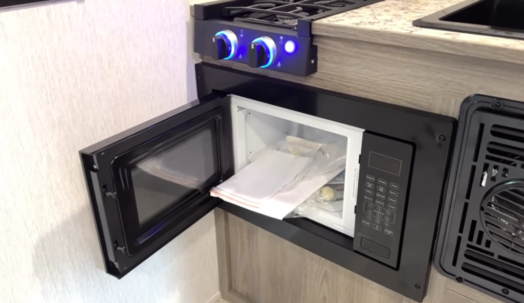Close-up view of an RV kitchenette featuring an open black microwave built into the cabinetry. Inside the microwave, there are factory-included accessories and manuals still in their plastic packaging. Above the microwave, a two-burner stovetop with illuminated blue control knobs is integrated into a light wood-finished countertop. To the right, a vent for air circulation is visible. The design is compact and modern, with neutral tones and space-efficient appliances.