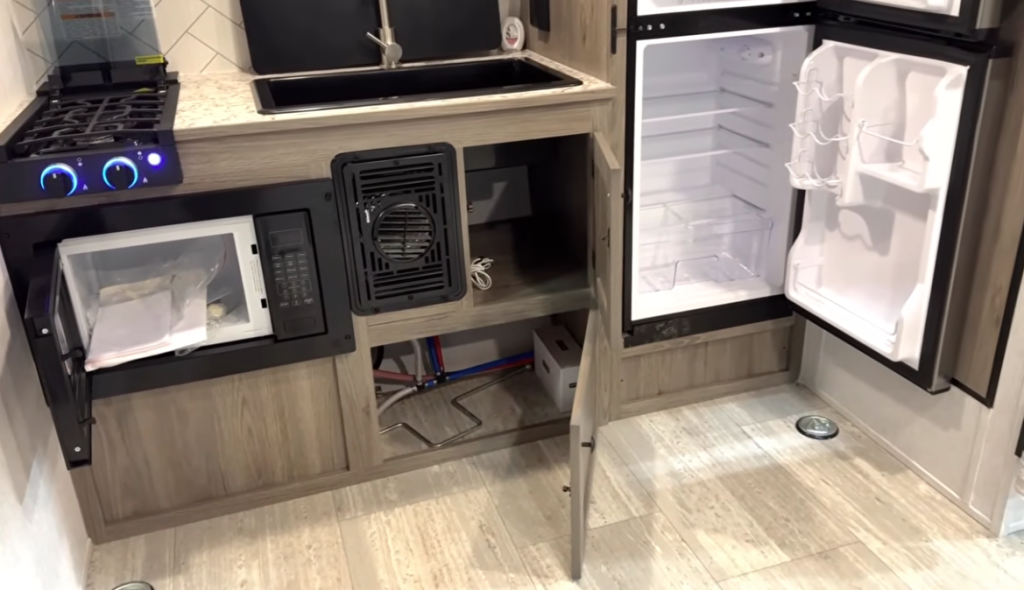 Interior view of an RV kitchen with opened appliances showing the functional components. On the left is a black microwave with its door open, showing a light-colored interior. The stovetop has blue illuminated control knobs. In the center is a black sink visible from above. To the right is a refrigerator with its door open, displaying the empty white interior shelving and door compartments. Below the sink and microwave area, a cabinet door is open revealing the plumbing and utility connections with visible pipes and wiring. The kitchen features light wood cabinetry and light wood-look flooring. The image provides a practical view of the RV's compact kitchen components and how the utilities are housed within the cabinetry. This appears to be from the same model shown in the previous image, with the white herringbone/chevron tile backsplash partially visible.
