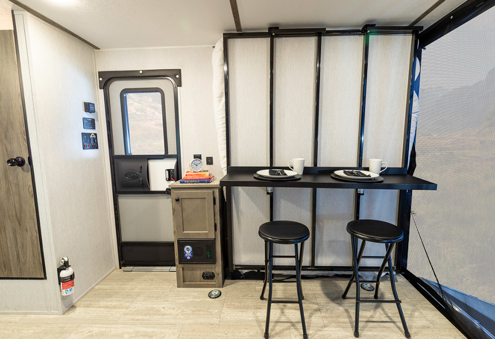 Interior of a compact travel trailer featuring a small black fold-down bar table with two matching black stools. The table is mounted against a reinforced wall near a large sliding glass door, allowing for natural light and an open feel. To the left, a small kitchenette area includes a light wood cabinet with a marble-look countertop and a built-in heater below. The flooring is light wood-look vinyl. In the background, a red carpet, gold stanchions, and a Pepsi vending machine indicate the RV is on display at an indoor event.