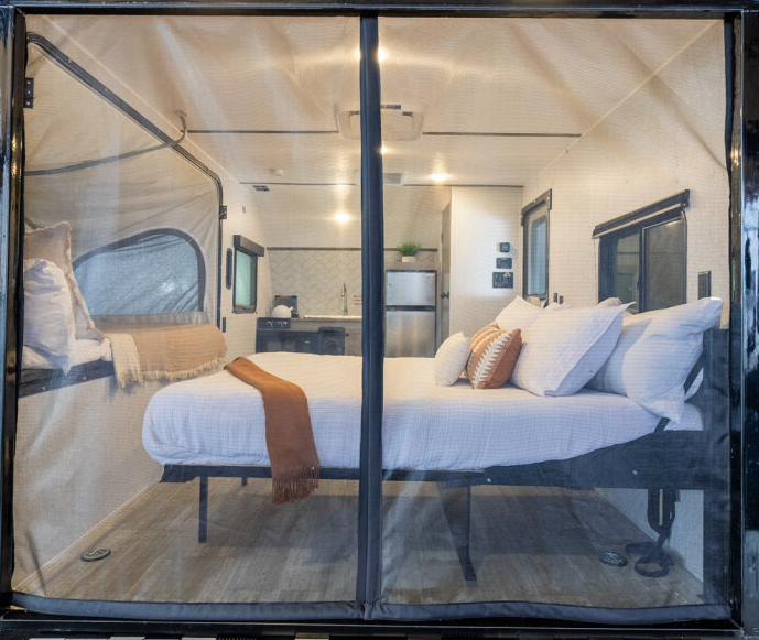 ALT text: View of an RV interior seen through a mesh screen door or window. The image shows a bedroom area with a platform bed featuring white bedding, multiple white pillows, and an orange or rust-colored throw blanket. Beyond the bedroom, parts of the kitchen area are visible with what appears to be a sink and refrigerator. The RV has light wood-look flooring and light-colored walls consistent with the modern design seen in previous images. The mesh screen creates a slight gauzy effect over the view but still allows visibility of the interior layout. This perspective appears to be from outside looking in through a screened entry door or large window, demonstrating how the RV looks when set up for camping with the screen providing ventilation while keeping insects out.