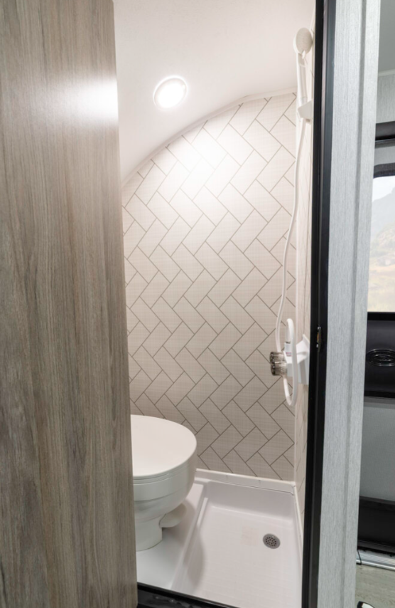 View of a compact RV bathroom through an open doorway. The small bathroom features a white toilet on the left side and a white shower pan or tray on the floor. The walls are covered with white subway tiles laid in a herringbone pattern, matching the kitchen backsplash seen in previous images of this model. The ceiling is curved, following the contour of the RV's roof, with a small round light fixture mounted in it. A white shower handle or control is visible on the wall. The doorway is framed with light wood-colored material that matches the interior design scheme of this RV model. The limited space demonstrates efficient use of the compact area while maintaining a clean, modern aesthetic. This bathroom appears to be from the same RV model shown in the most recent kitchen images, featuring the coordinated herringbone tile pattern throughout.