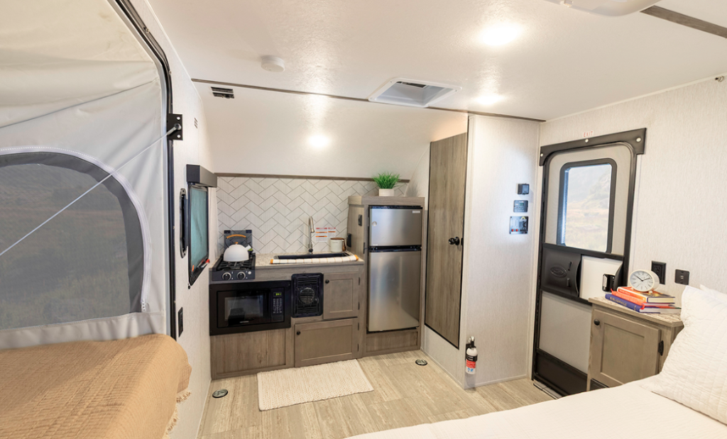  Interior view of the compact camper trailer showing the complete main living area. On the left is part of the pop-out sleeping area with a beige/cream bedspread, enclosed in canvas with mesh windows. The kitchen area occupies the back wall with the same features seen in previous images: stainless steel refrigerator/freezer, black microwave, two-burner stovetop, sink with faucet, and the distinctive white herringbone tile backsplash. Light wood-tone cabinetry is used throughout. A wooden door (likely leading to the bathroom) is visible beside the refrigerator. On the right side is the main entrance door with a window, and next to it is a small counter or desk area with some books and a clock. A fire extinguisher is mounted on the wall near the entrance. The floor has light wood-look laminate with a small cream rug in front of the kitchen. The ceiling features recessed lighting and a vent. This wide-angle view shows how the compact trailer efficiently organizes the kitchen, sleeping, and living spaces into a cohesive, functional design.