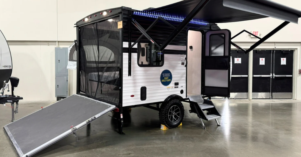 Exterior view of a compact travel trailer or micro camper displayed in what appears to be a showroom or exhibition hall. The camper is primarily white with black accents and features a distinctive boxy design. It has several notable features including: a black door on the right side with steps leading up to it, a large mesh-screened pop-out section on the left side (likely the expandable sleeping area seen in previous interior shots), a ramp or loading platform extending from the left side, a black awning extended from the top, and blue LED lighting along the edge of the awning. The trailer is equipped with off-road tires and appears to be designed for rugged use. It's sitting on a concrete floor in an indoor display space with exit doors visible in the background. A small blue award or certification emblem is attached to the side of the trailer. This appears to be a compact, feature-rich camper designed to maximize space through expandable sections while maintaining mobility and off-road capability.