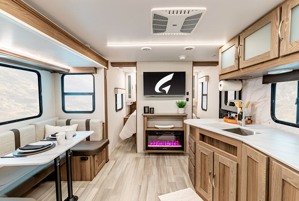 ALT text: Interior view of a modern RV or travel trailer featuring a spacious living area with light wood finishes. The left side shows a dining area with bench seating and a table set with plates and cups. On the right is a kitchen counter with sink and faucet. The center features a flat-screen TV mounted above an electric fireplace with purple-pink flames. The design includes white countertops, wood cabinetry throughout, large windows, ceiling vents, and comfortable seating. The space has a clean, contemporary aesthetic with warm lighting.