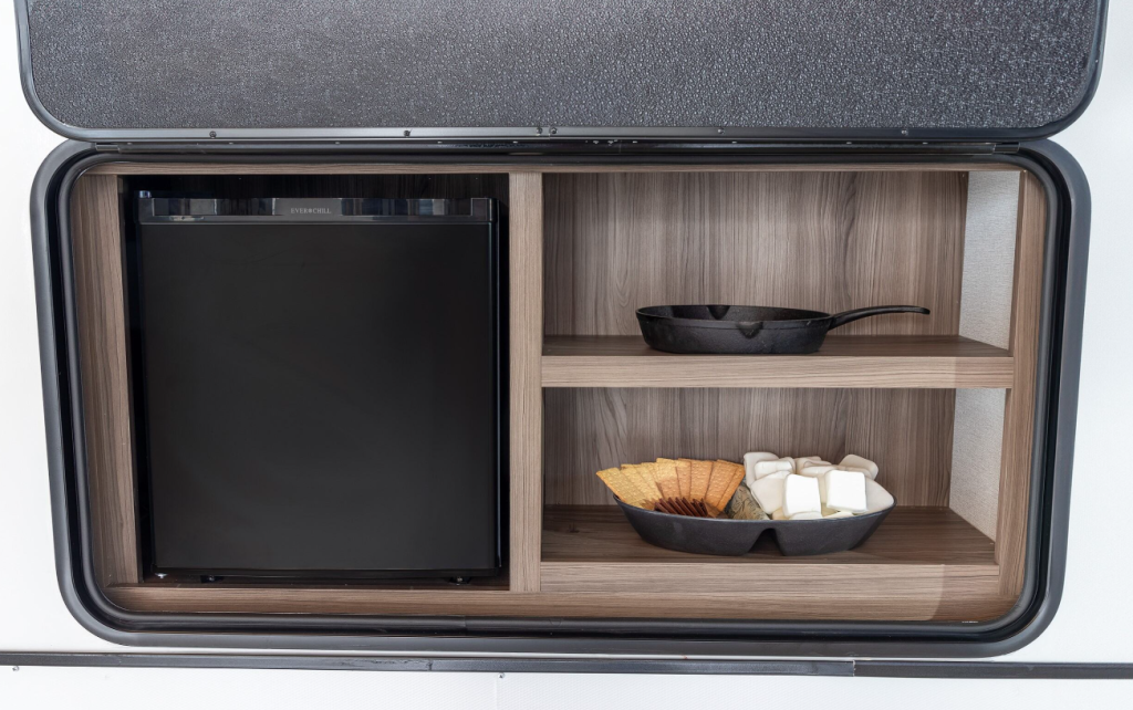 An exterior view of an RV outdoor kitchen compartment accessed through a side hatch. The compartment is built into the side of the RV with a black frame around the opening. Inside the compartment, there's a small black refrigerator or cooler on the left side. On the right side are two wooden shelves matching the light wood finish seen throughout the RV's interior. The top shelf contains a black cast iron or non-stick frying pan with handle. The bottom shelf holds a black serving dish containing graham crackers or similar crackers and marshmallows, likely for making s'mores. This outdoor kitchen area allows for cooking and food preparation outside the RV while camping.