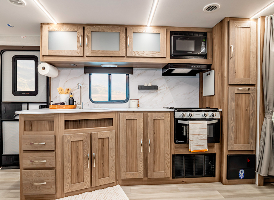  Interior view of an RV kitchen area featuring a complete kitchen setup with light wood cabinetry throughout. The kitchen includes upper cabinets with frosted glass panels, a black microwave and range hood mounted above a cooktop/oven. The countertop is white with a marble-like pattern and includes a sink with a modern faucet. There's a small window above the backsplash, which has the same marble-like finish as the counter. Lower cabinetry includes multiple drawers and cabinet doors with silver handles. A paper towel holder is mounted on the left side of the kitchen area. The same striped kitchen towel seen in previous images hangs from the oven handle. A small cream-colored rug lies on the light wood-look flooring. The right side shows tall cabinet storage and what appears to be the edge of a curtain. The ceiling features recessed lighting and air vents.