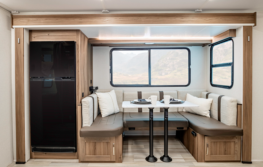  Interior view of an RV or travel trailer dining area. The space features a U-shaped dining booth with cream-colored cushions and brown accents, arranged around a white table mounted on black pedestals. White pillows rest on the bench seating. Large windows with black frames provide views of mountainous terrain outside. To the left is a black refrigerator or storage cabinet integrated into the wooden cabinetry. The dining area is framed by light wood paneling that matches the lower storage cabinets beneath the seating. The flooring appears to be a light vinyl or laminate designed to resemble wood.