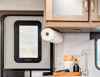 Close-up view of a section of the RV kitchen area. The image shows a small window with a black frame on the left side, likely providing ventilation or light to the kitchen space. Next to the window is a paper towel holder mounted under the upper cabinets with a roll of white paper towels. The kitchen counter is visible with part of a wooden utensil holder containing what appear to be wooden spoons or spatulas. Also visible is a portion of the sink area with a modern stainless steel faucet. The backsplash features the same white marble-like pattern seen in previous images, and the upper cabinetry with light wood frames and frosted glass inserts is partially visible at the top of the frame. The image captures a small but functional detail of the RV's kitchen organization.