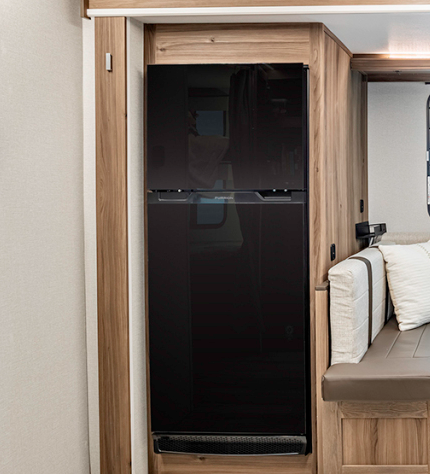 Close-up view of a black refrigerator in an RV. The refrigerator has a sleek, modern design with a two-door configuration (top and bottom) and is built into light wood cabinetry that matches the overall interior design of the RV. The refrigerator has a glossy black finish with horizontal handles separating the upper and lower compartments. To the right side of the image, a portion of the RV's dining area is visible, showing cream-colored bench seating with white pillows and a brown seat base with wooden trim. The refrigerator is positioned near the dining area, maximizing the efficient use of space typical in RV design.