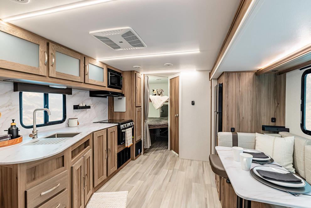 Interior view of an RV showing a full-length perspective of the main living area. On the left is the kitchen with light wood cabinetry, white countertops with a marble-like backsplash, sink with modern faucet, and built-in appliances including a microwave and stovetop/oven. The right side shows the dining area with bench seating and a table set with plates and cups. The central hallway leads to what appears to be a bathroom or bedroom area visible through a partially open door. The RV features consistent design elements throughout, including light wood cabinetry with silver hardware, white walls, light wood-look flooring, frosted glass cabinet fronts, recessed lighting, and ceiling vents. Windows with black frames are visible on both sides of the space. A small cream-colored rug is placed on the kitchen floor.