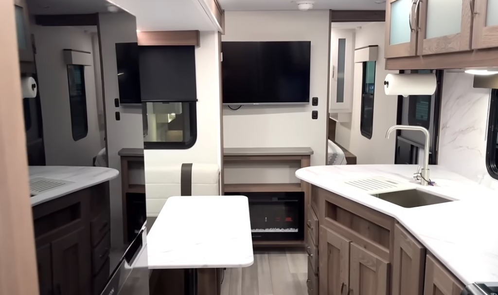  Interior view of the RV showing the main living area from a different angle. In the foreground is the dining table with a white surface. On the right is the kitchen counter with sink and white countertops featuring a marble-like pattern. The backsplash matches the countertop material. Light wood cabinetry is seen throughout. In the center background is an electric fireplace set into the wall beneath what appears to be mounted screens or TVs with black surfaces (possibly turned off). The cream-colored bench seating with brown accents is partially visible around the sides. Windows with black frames can be seen on the walls. This view shows how the compact living space is arranged, with the kitchen, dining, and entertainment areas all visible within the same open concept area. The consistent design elements of light wood, white surfaces, and black accents are maintained throughout the space.
