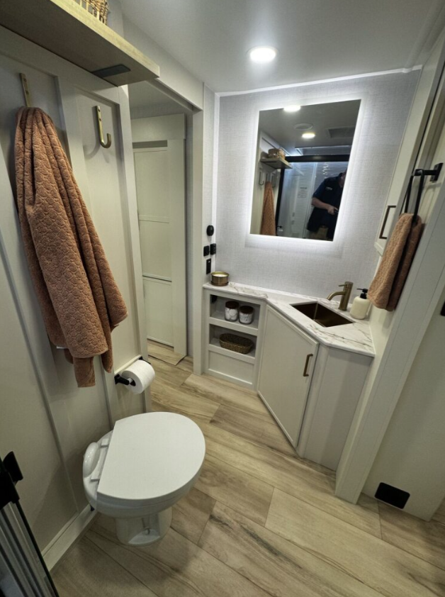 ALT text: A wider view of the Brinkley RV bathroom taken from a different angle, showing more of its features. The white toilet is on the left with a tan towel hanging from a hook on the wall above it. To the right is a compact vanity area with a small sink, a brass/gold faucet, and white cabinetry with open shelving containing decorative items. Above the vanity is a mirror with integrated LED lighting around its perimeter. In the back appears to be a door, possibly leading to a shower area or closet. The bathroom has the same light wood laminate flooring as the rest of the RV, white walls, and a white ceiling with recessed lighting. Another tan towel is visible hanging on the door to the right. 