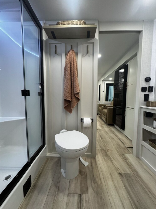 ALT text: Interior view of the bathroom in the Brinkley RV. The bathroom features a white toilet positioned against a white wall panel with decorative vertical grooves. Above the toilet is a small shelf holding a wicker basket, and wall hooks with a tan towel hanging. A toilet paper holder is mounted on the wall. To the left is a shower enclosure with a glass door and black frame. Looking out from the bathroom toward the main living area, the tan leather seating and part of the kitchen are visible. The bathroom continues the same light wood laminate flooring seen throughout the RV. Control panels are visible on the wall to the right. 