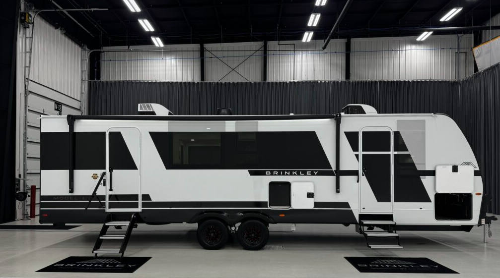 Exterior view of a Brinkley travel trailer displayed in what appears to be a showroom or manufacturing facility. The travel trailer features a modern, sleek design with a distinctive white and black exterior color scheme with geometric pattern elements. The side of the trailer prominently displays the "BRINKLEY" brand name. The RV includes multiple windows, an entry door with steps extended down, and appears to be a large model sitting on dual axles with black wheels. The showroom has a white floor with "BRINKLEY" branded mats positioned near the trailer, white metal walls, dark curtains in the background, and fluorescent lighting in the ceiling. 