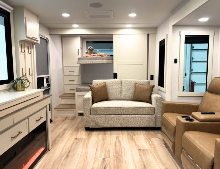 Interior view of the Brinkley RV showing a modern, apartment-like living space. The image features a bright, open living area with light wood laminate flooring and white walls and cabinetry. In the foreground right are two tan leather recliners. Across from them is a light gray loveseat with two brown accent pillows. Behind the loveseat is an elevated sleeping area or bunk, accessed by small stairs with built-in storage drawers. A golden retriever dog is visible lying on the elevated bunk/bed. The left side of the image shows part of the kitchen area with white cabinetry, drawers with silver handles, and what appears to be appliances. The ceiling has recessed lighting, and there's a small houseplant visible on a counter or shelf. Doorways to other areas of the RV are visible in the background.
