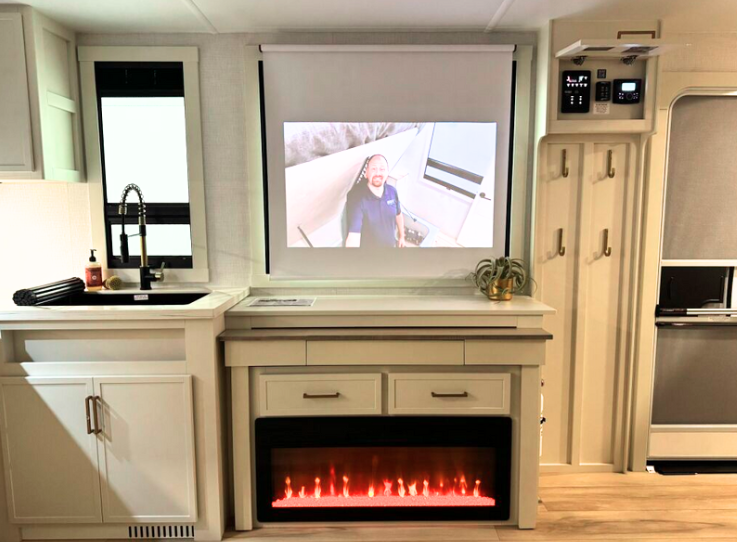 Interior view of the Brinkley RV entertainment wall and kitchen area. The center of the image features a built-in electric fireplace with glowing red flames in a black housing, integrated into light-colored cabinetry with drawers above it. Directly above the fireplace is a pull-down projection screen showing an image of what appears to be the same bearded man (likely the "RV Nerd") seen in previous images. To the left is part of the kitchen with a sink featuring a black faucet, white countertops, and white cabinetry with silver handles. On the right side are coat hooks and what looks like control panels, possibly for RV systems. A small houseplant is visible on the counter next to the projection screen. 