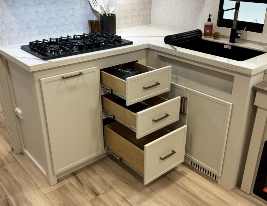  Close-up view of the kitchen corner in the Brinkley RV, focusing on storage features. The image shows the L-shaped kitchen counter with a black gas cooktop on the left and a black sink with black faucet on the right. Three drawers are pulled open beneath the counter between the cooking and sink areas, revealing wooden drawer interiors with what appear to be black-labeled dividers or organizers. The cabinetry is cream/off-white with brushed metal handles, and the countertop is white. The backsplash features white subway tiles. On the countertop behind the cooktop is a black container holding kitchen utensils. Near the sink are decorative items including what looks like a small golden/brass decorative object and a soap dispenser. The white countertop extends around the corner, and cabinet doors are visible beneath the sink area. 