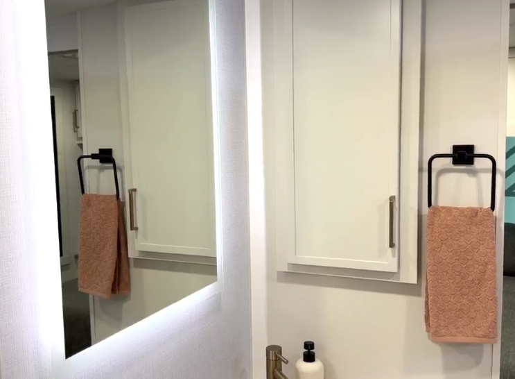  A bathroom detail in the Brinkley RV showing a mirror with integrated LED backlight, creating a glowing perimeter effect against the white wall. The mirror reflects a towel ring on the opposite wall holding a peachy/tan hand towel. On the right side of the image is a matching towel ring with another peachy/tan hand towel. White cabinet doors with small silver handles are visible on the wall. The bottom of the image shows part of the brass faucet and white soap dispenser seen in the previous close-up of the sink area. 