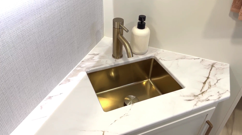 Close-up view of the bathroom sink in the Brinkley RV, featuring premium design elements. The image shows a corner-mounted vanity with a white marble-look countertop featuring subtle gray veining. The sink is a square, undermount design in a gold/brass finish that coordinates with the matching brass faucet. A white ribbed soap dispenser with a black pump sits on the counter. The backsplash appears to be a textured light gray tile. This detailed view highlights the upscale finishes used in the bathroom, with the warm metallic tones of the brass fixtures contrasting beautifully with the cool white marble-look surface. 