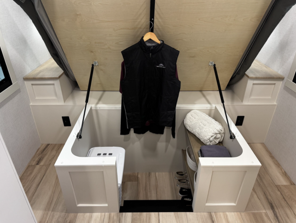 An ingenious storage solution in the Brinkley RV bedroom showing a dual-purpose closet and storage area. The image reveals a walk-in closet space with a black Brinkley-branded vest hanging on a wooden hanger in the center. On either side are white cabinet structures with tops that are hinged upward and supported by gas struts, revealing deep storage compartments. The left compartment contains what appears to be a laundry basket or container, while the right side contains folded linens or clothing and possibly shoes at the bottom. The area has light wood paneling on the ceiling/upper walls and the same light wood laminate flooring seen throughout the RV. 
