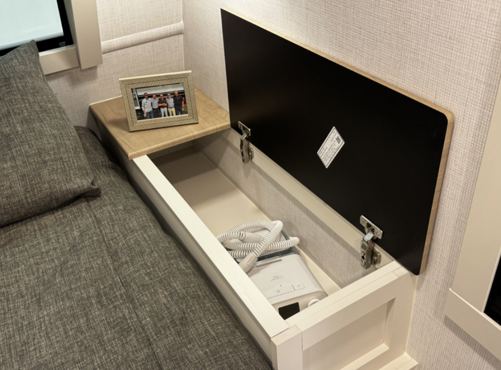 Close-up view of a nightstand in the Brinkley RV bedroom with clever hidden storage. The image shows a bedside table with a wooden top where a framed photograph of a group of people is displayed. The top of the nightstand is hinged and open, revealing a hidden compartment beneath. Inside this compartment is what appears to be a CPAP machine (continuous positive airway pressure device used for sleep apnea) with its white hose visible. The compartment allows for convenient storage of the medical device while keeping it accessible for nighttime use. The black underside of the lid has a small white information sticker. On the left side of the image is part of the gray bed with matching gray pillow. The wall has a subtle textured light gray covering.
