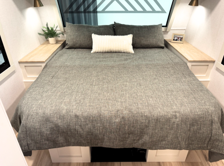 View of the bedroom area in the Brinkley RV featuring a queen or king-sized bed with a gray textured bedspread/comforter and three pillows - two larger gray pillows and one smaller white ribbed accent pillow in the center. The bed is flanked by built-in nightstands with light wood tops and white bases. Each nightstand has a wall-mounted brass/gold sconce light above it. On the left nightstand is a small potted plant, and the right nightstand has what appears to be a small picture frame. Above the bed is a decorative headboard or wall feature with geometric patterns visible through glass. Beneath the bed appears to be storage drawers or compartments with white cabinet faces.