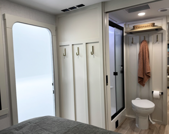 View of the bedroom and part of the bathroom in the Brinkley RV. On the left side is a corner of the bed with the gray bedspread visible. The center of the image shows a white wall panel with built-in coat hooks or hangers. To the left of these hooks is what appears to be an RV entry door with a window. On the right side, through an open doorway, is a glimpse into the bathroom showing the white toilet, a peach/tan towel hanging on a hook, and a basket on a shelf above. The doorway to the bathroom has a sliding glass door with a black frame. 