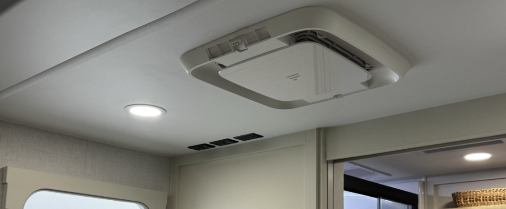 A close-up view of the ceiling in the Brinkley RV showing a modern air conditioning unit. The image displays a low-profile, square white air conditioning vent or unit mounted flush with the white ceiling. Next to it is a circular LED recessed light fixture. Some air vents or registers are visible on the wall below the ceiling. The upper portion of what appears to be a doorway or opening to another area of the RV is visible in the lower right corner. A small portion of what might be a window frame can be seen in the lower left. 