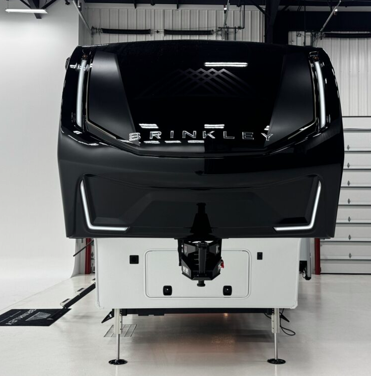 Front view of a Brinkley fifth wheel RV showcasing its sleek black aerodynamic cap, illuminated LED accent lights, and bold BRINKLEY branding in a modern service bay setting.