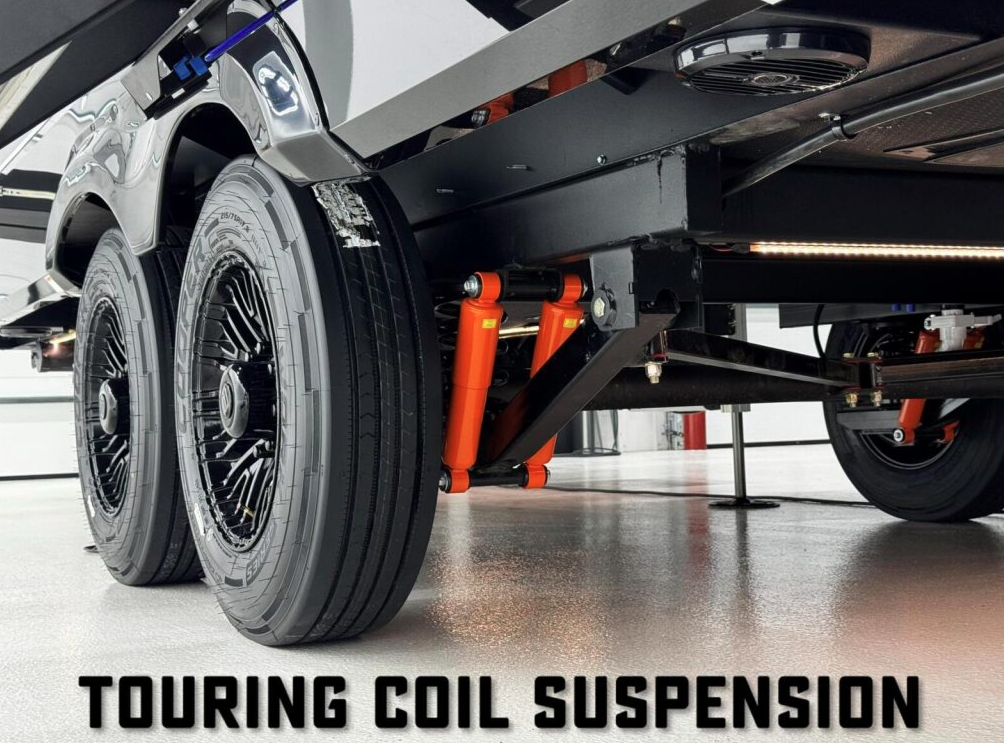 Close-up view of a Brinkley RV’s touring coil suspension system, showcasing dual black alloy wheels, heavy-duty orange shock absorbers, and coil components mounted on a clean shop floor.