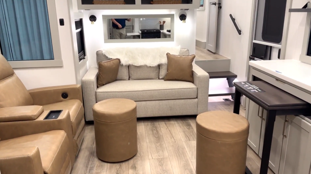 Interior view of a modern RV living space featuring tan leather recliners, a beige sofa with decorative pillows, two ottomans, and a compact work desk, all set against light wood flooring and bright white cabinetry.