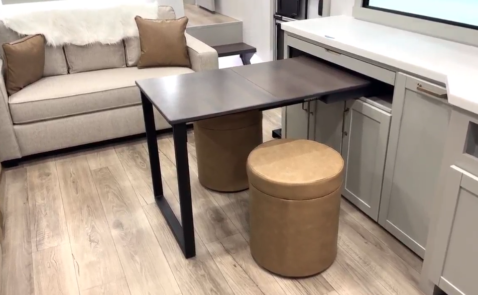Convertible RV workstation with a pull-out dark wood table and two tan leather ottoman stools, positioned in front of light gray cabinetry and across from a beige sleeper sofa on wood-style flooring.








