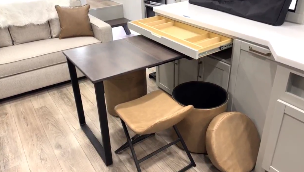 Compact RV dining and workstation area featuring a dark folding table, a leather padded stool, storage ottoman with removed lid, extended silverware drawer, and a nearby beige sofa, all set on wood-look vinyl flooring.