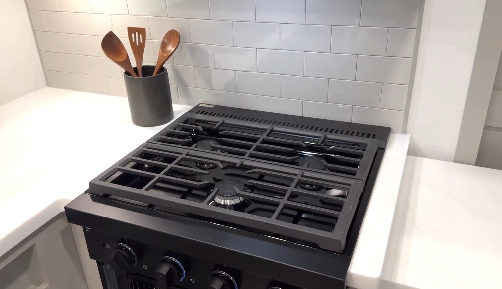 Close-up of a modern black gas stovetop with three burners and heavy-duty grates in an RV kitchen, set against a white tile backsplash and flanked by white solid-surface countertops; a black utensil holder with wooden cooking tools sits in the corner.