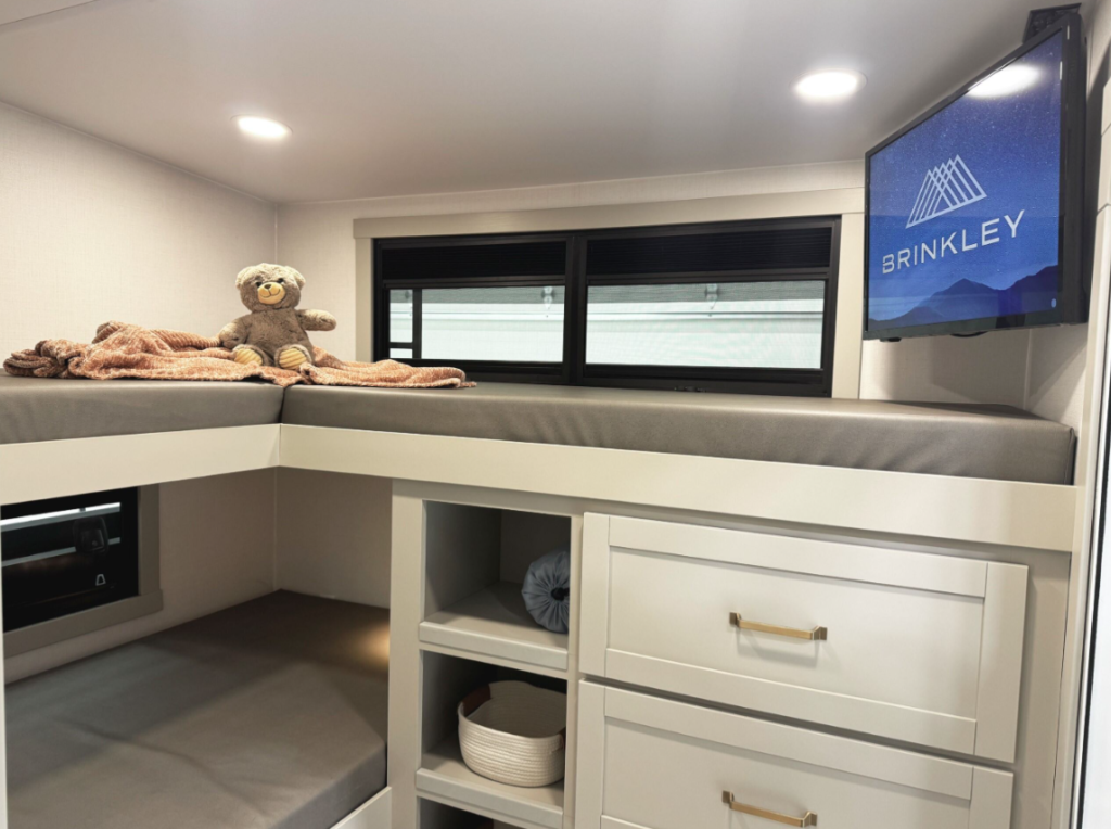  Bright and modern bunkhouse in a Brinkley RV featuring an L-shaped upper and lower bunk with gray cushions, built-in drawers and shelving, dual windows with black-out shades, a wall-mounted TV displaying the Brinkley logo, and a plush teddy bear on a cozy blanket.